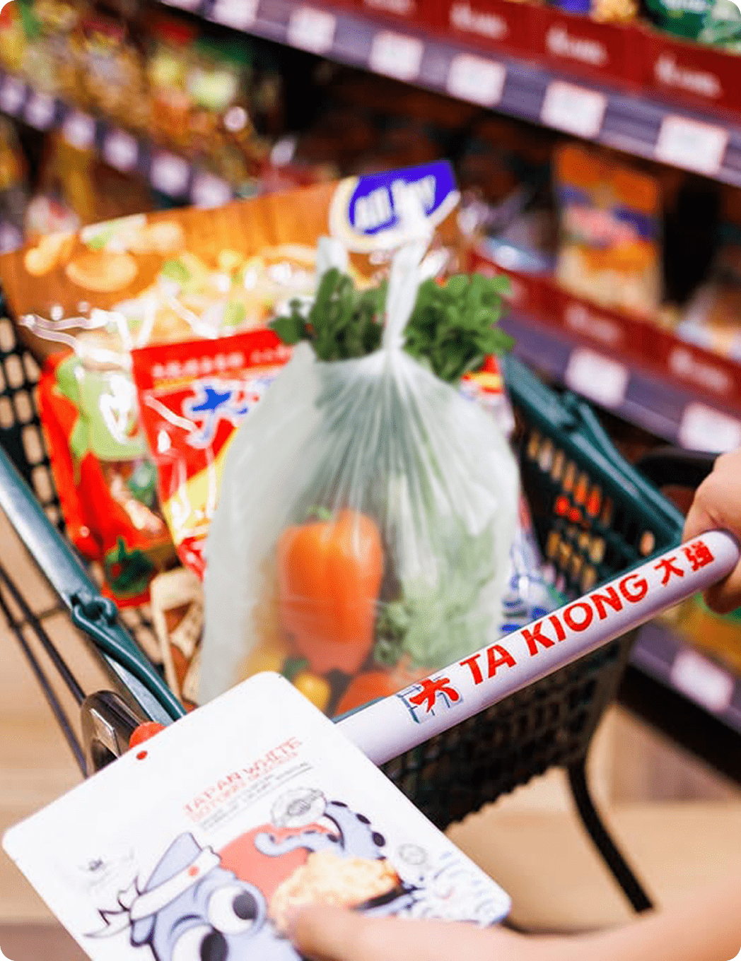 Shopping cart in grocery aisle with colorful goods. Hand holds "Japan White" pouch; handle reads "Ta Kiong."