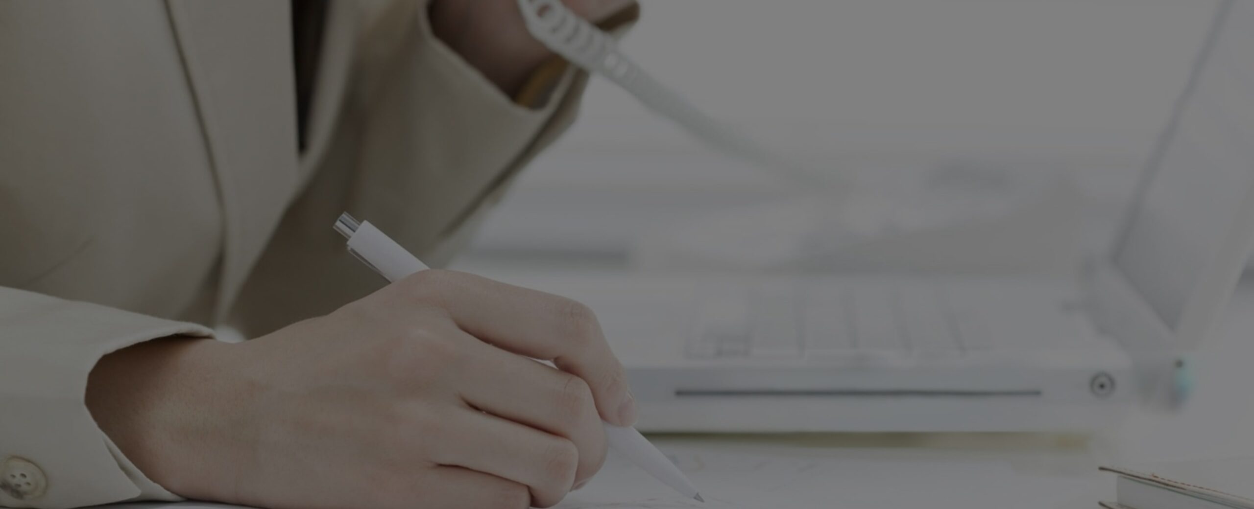 Close-up of a suited person using a phone and pen near a laptop in a focused, professional setting.